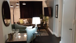 Un salon avec un canapé, une table basse et une lampe. Un téléviseur est fixé au mur et une porte donne accès à une autre pièce.