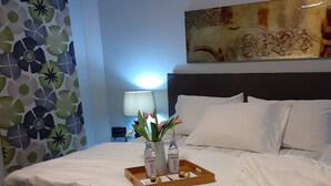 Un lit bien fait, avec du linge de lit blanc, un plateau avec des bouteilles d’eau et un vase de fleurs, et un tableau accroché au mur.