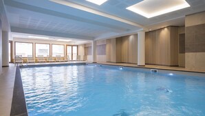 Une piscine intérieure à l’eau claire, avec un sol carrelé et une pièce lumineuse grâce à de grandes fenêtres et des fauteuils.
