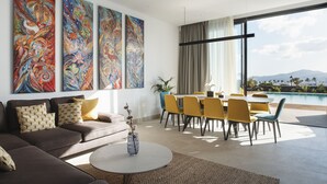 A modern living room with a sofa, coffee table, and dining area. There are three large paintings on the wall and a view of a pool and mountains through large windows.