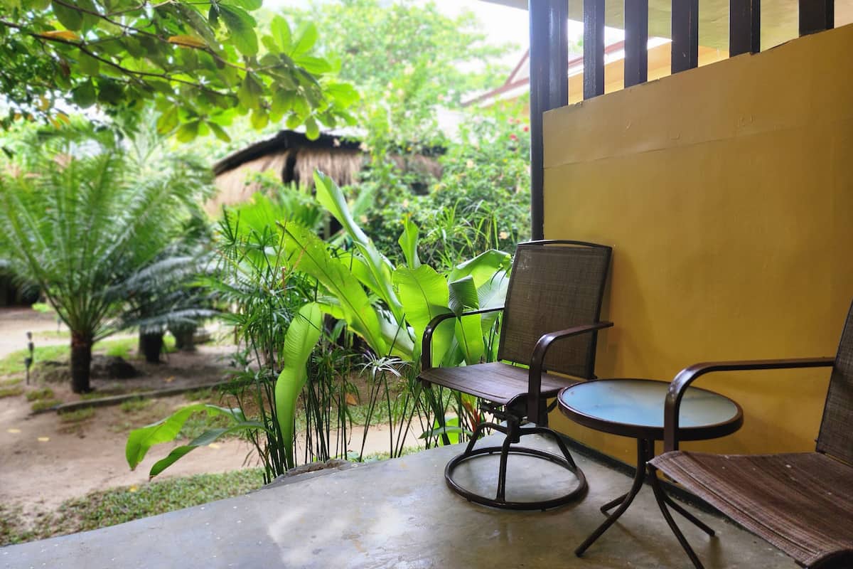 Eine Terrasse mit Stuhl und Tisch, umgeben von üppiger Vegetation und einem Reetdach im Hintergrund.