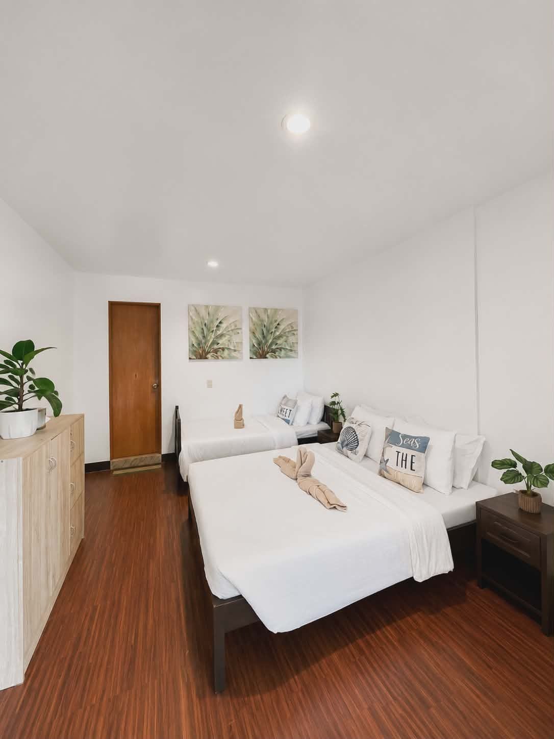A hotel room with two beds, a wooden wardrobe, a small table, and a potted plant.