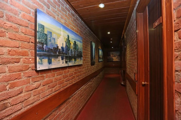 A corridor with brick walls and wooden doors, leading to a room with a red carpet.