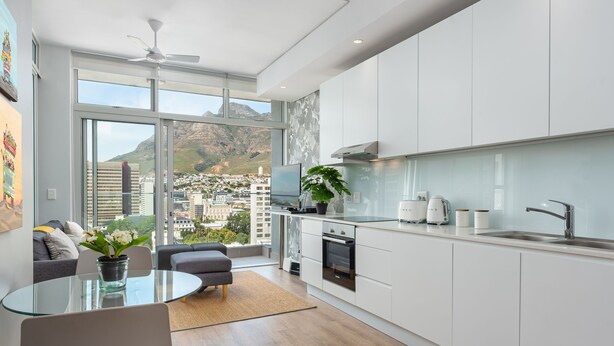 A modern kitchen with white cabinets, a glass dining table, and a view of a cityscape through large windows. - The Sentinel 1310 (Cape Town)