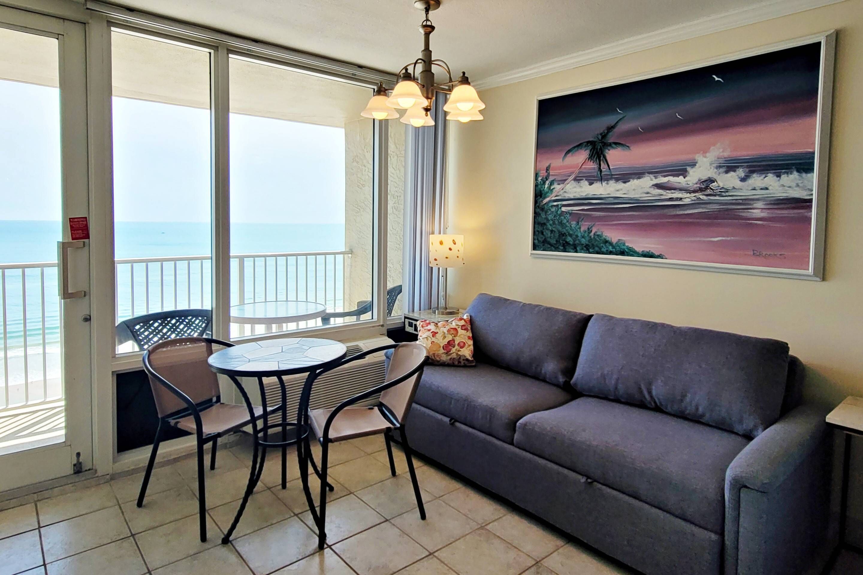 A balcony with a view of the sea, a gray sofa, a round table with two chairs, and a painting of a beach scene.