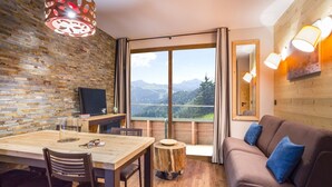A cozy room with a wooden dining table, chairs, a sofa with blue cushions, a TV, and a large window offering a mountain view. - Modern Comfort by the Slopes (Tignes 1800)