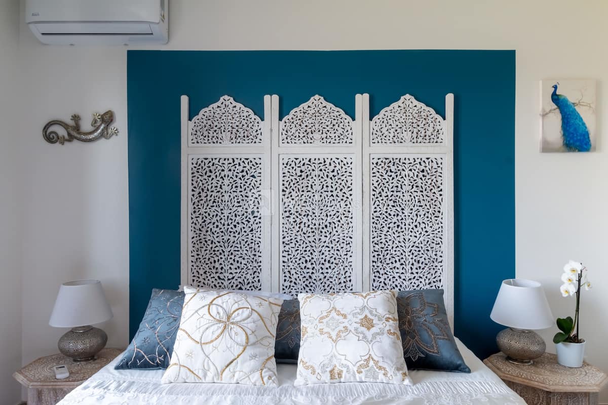A bedroom with a bed, decorative pillows, a nightstand with a lamp, and a wall-mounted air conditioner.
