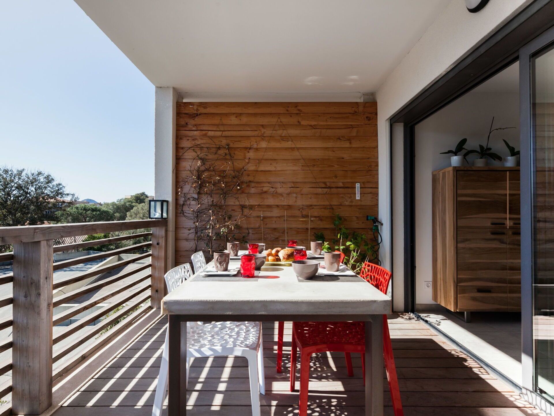 Een balkon met een houten tafel en stoelen, uitzicht op zee en een modern gebouw met glazen deuren.