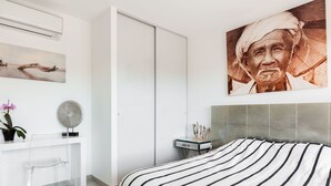 A modern bedroom with a large bed, a white desk, a small table, and a wall-mounted air conditioner. - Cosy apartment for 2 guests with WIFI, pool, A/C, TV and terrace (Porto-vecchio)