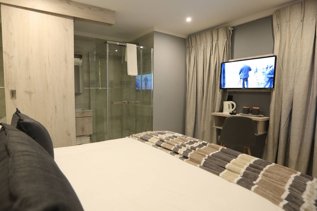 A hotel room with a bed, a TV, a shower area, and a desk with a kettle.
