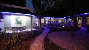 En hotellentrén med en skylt som visar texten ”CASCADES GUEST LODGE”, ett utomhusområde med sittplatser och en vackert anlagd trädgård.