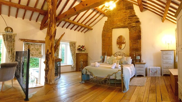 A bedroom with a stone fireplace, wooden beams, a bed with pillows, a nightstand, and a dresser. - Romantic, quirky, unique cottage, big garden, free parking, pet friendly max 2 (Beckermet)