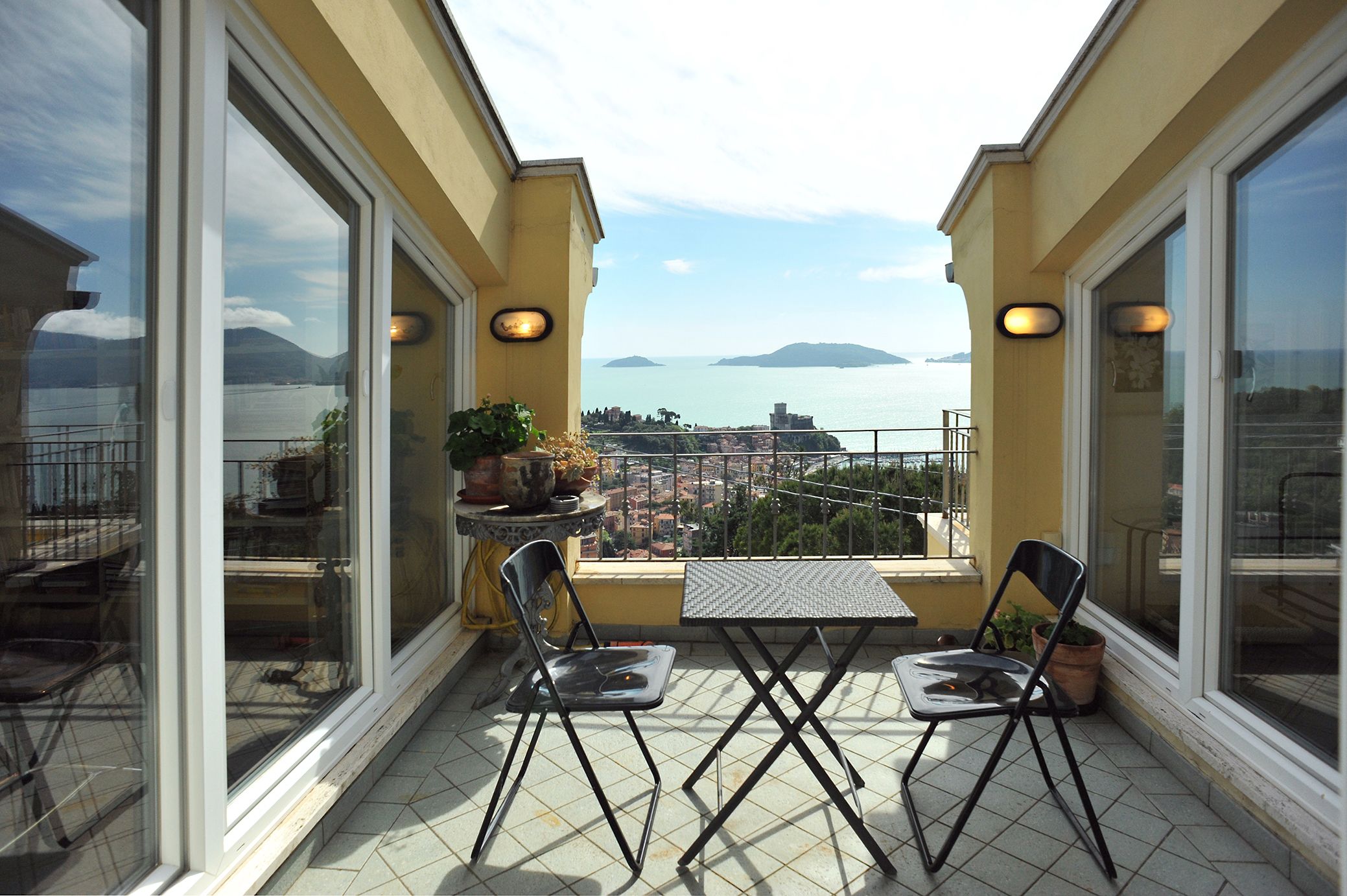 Un balcone con due sedie e un tavolo, affaccia su una città costiera con montagne sullo sfondo.