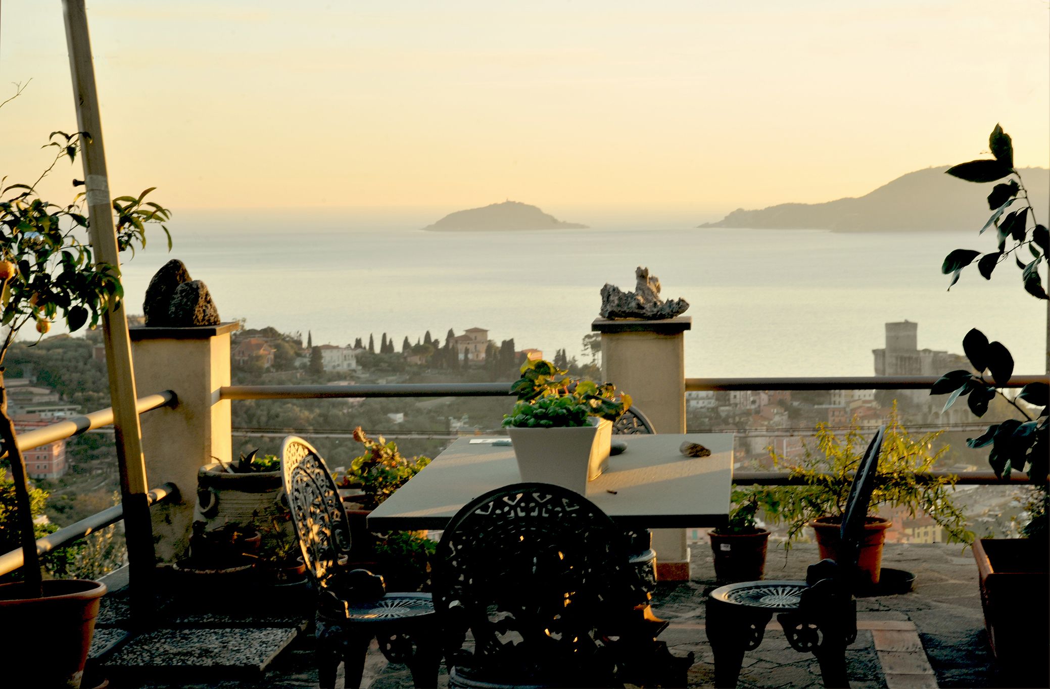 Un balcone con tavolo e sedie, piante in vaso e vista sul mare e sulle colline in lontananza.
