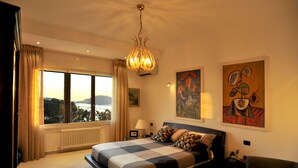 A bedroom with a large bed, a view of the outdoors, and two paintings on the wall.