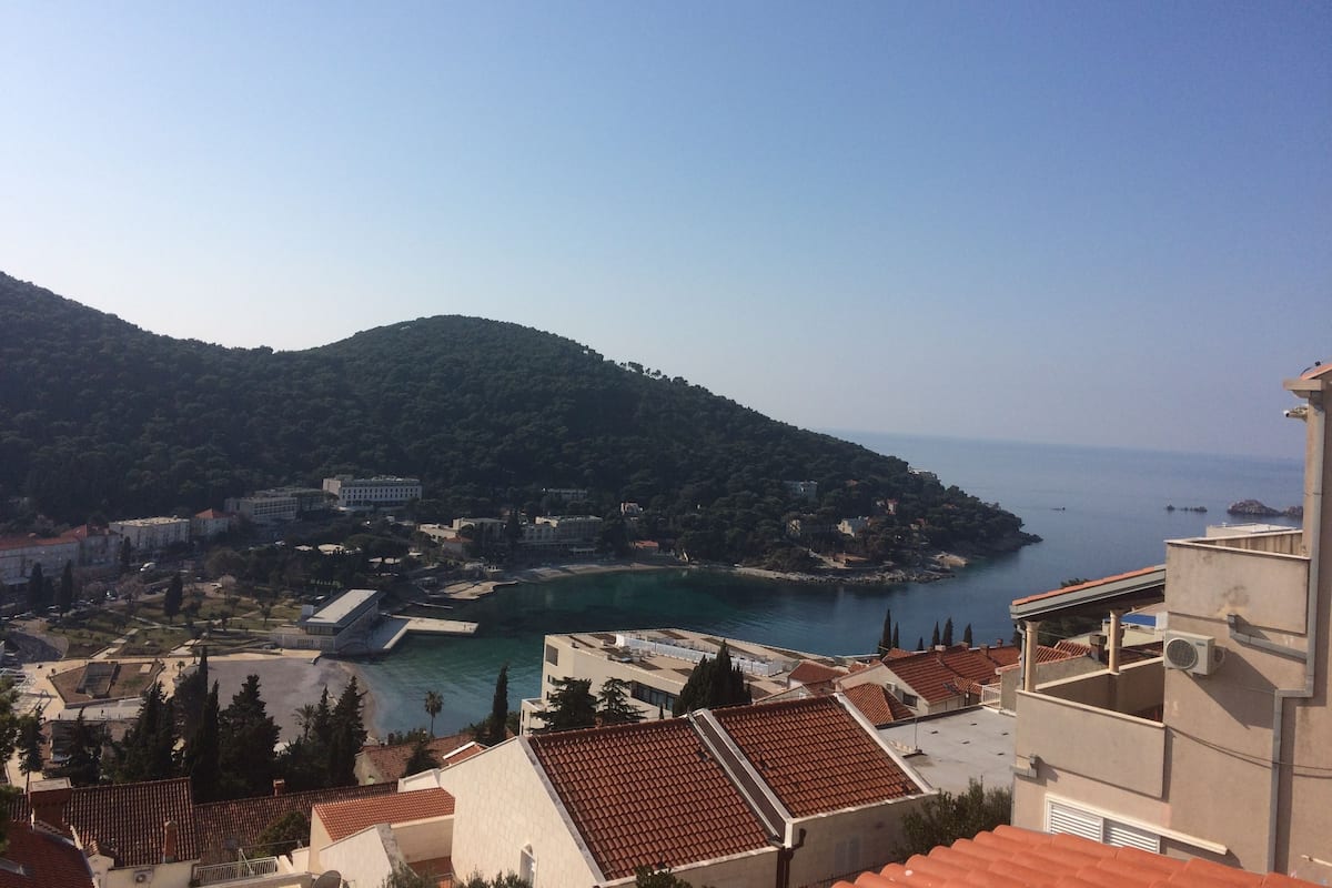 A coastal town with terracotta-roofed buildings, a marina, and a hillside with dense greenery.