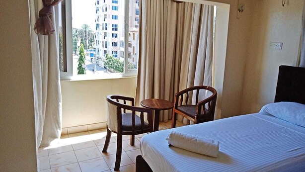 A hotel room with a bed, a small table, two chairs, a window with curtains, and a view of buildings outside. - Regency Inn (Mombasa)