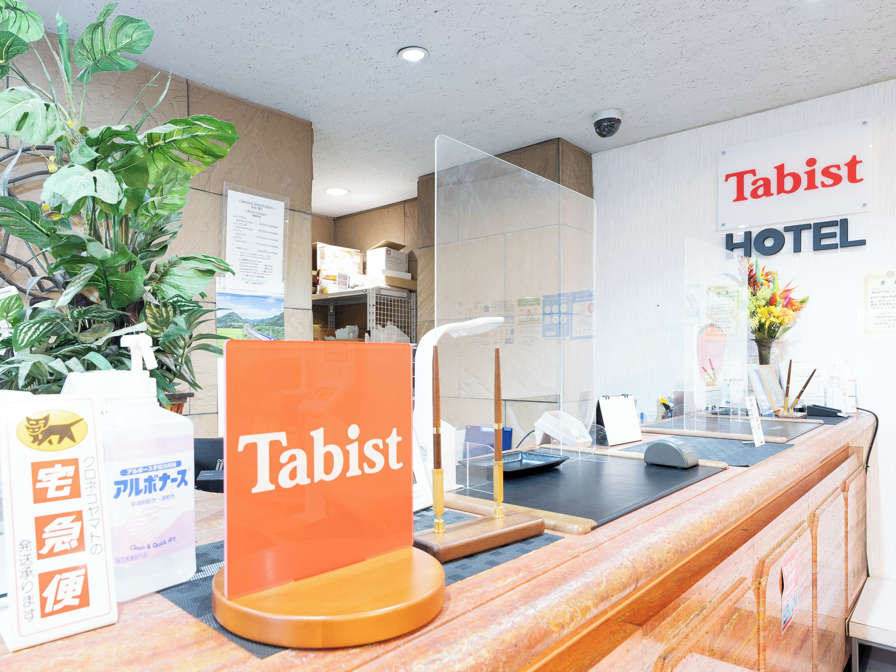 A hotel reception area with a sign reading "Tabist HOTEL," a reception desk, a security camera, and a potted plant.