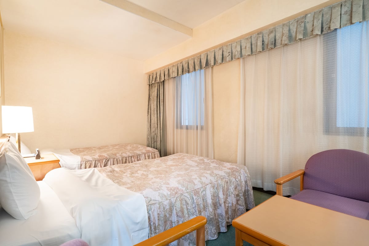 A hotel room with two beds, a chair, a table, and a lamp.