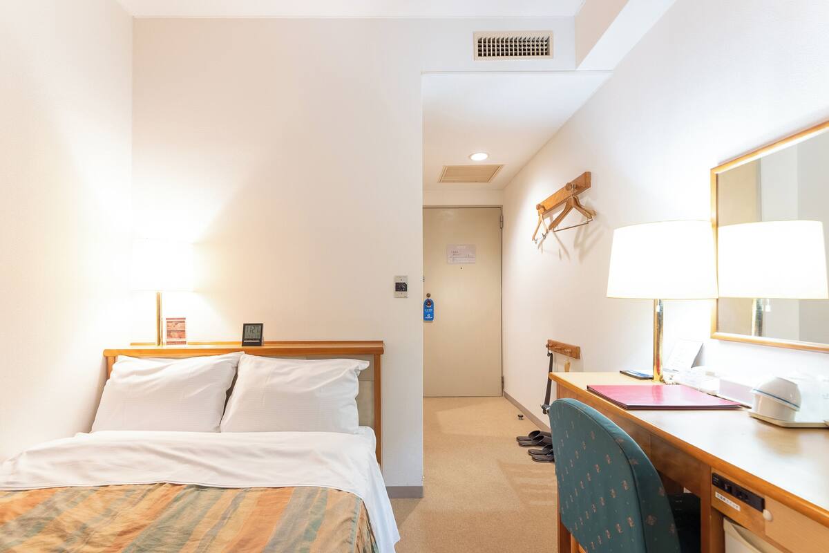 A hotel room with a bed, a desk with a chair, a lamp, a mirror, and a door leading to another room.