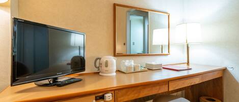 A hotel room with a wooden desk, a television, a kettle, a mirror, a lamp, a trash can, and a small stool.
