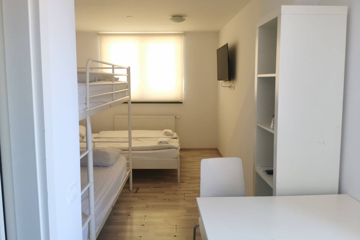 A small, well-lit room with bunk beds, a single bed, a desk, and a TV mounted on the wall.