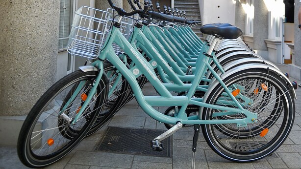 Uma fileira de bicicletas azuis estacionadas em frente a um prédio com paredes brancas e telhado telhado.