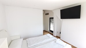 Quarto moderno e minimalista com duas camas, uma TV de tela plana na parede e uma janela grande que oferece luz natural.