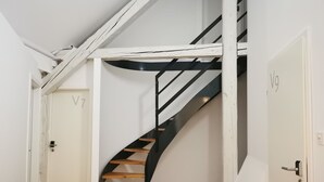 Uma escadaria em espiral com degraus de madeira e grades pretas, que leva a uma porta com a inscrição "V7".
