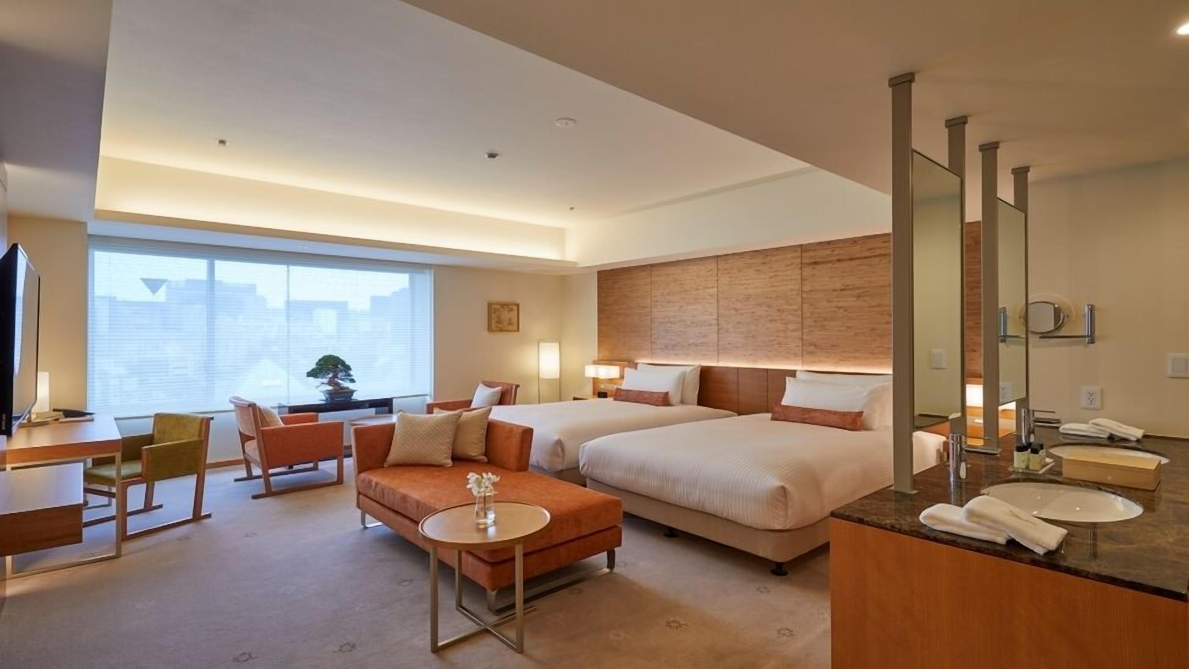 A hotel room with two beds, a desk, a sofa, a small table, and a bathroom with a mirror and sink.