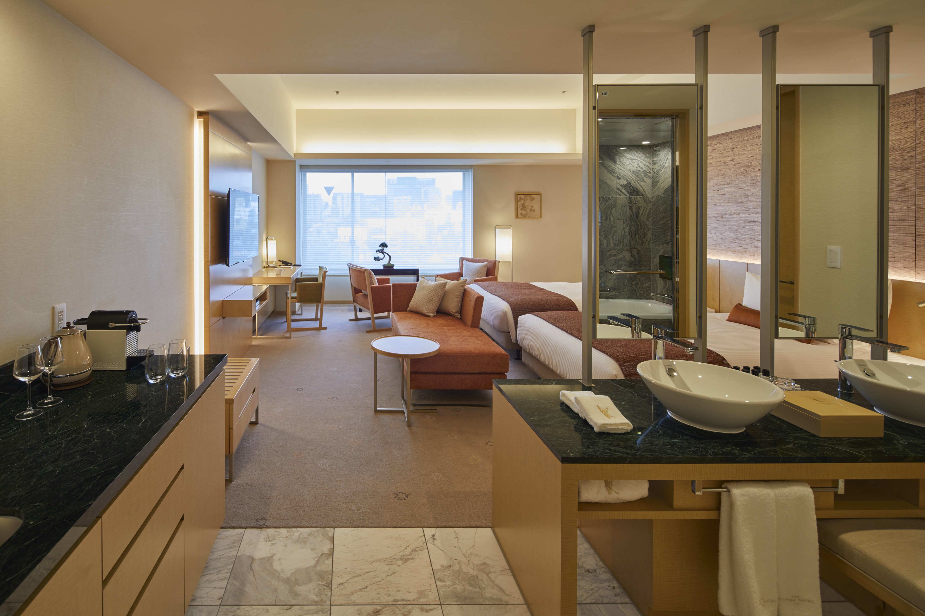 Quarto de hotel moderno com uma cama grande, um espaço de estar, uma mesa de jantar e banheiro com um espelho amplo e duas pias.