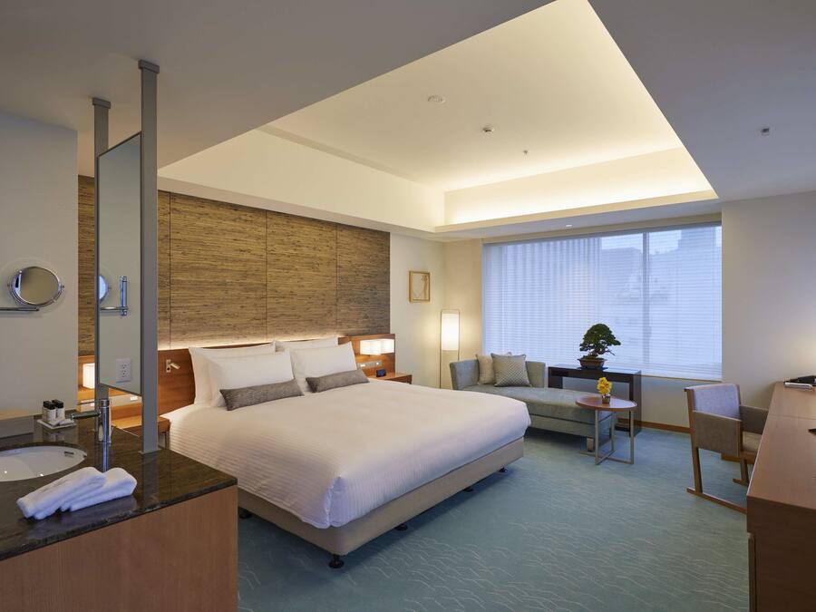A modern hotel room with a large bed, a sitting area with a sofa and a small table, a desk with a chair, a TV, and a large window with blinds.