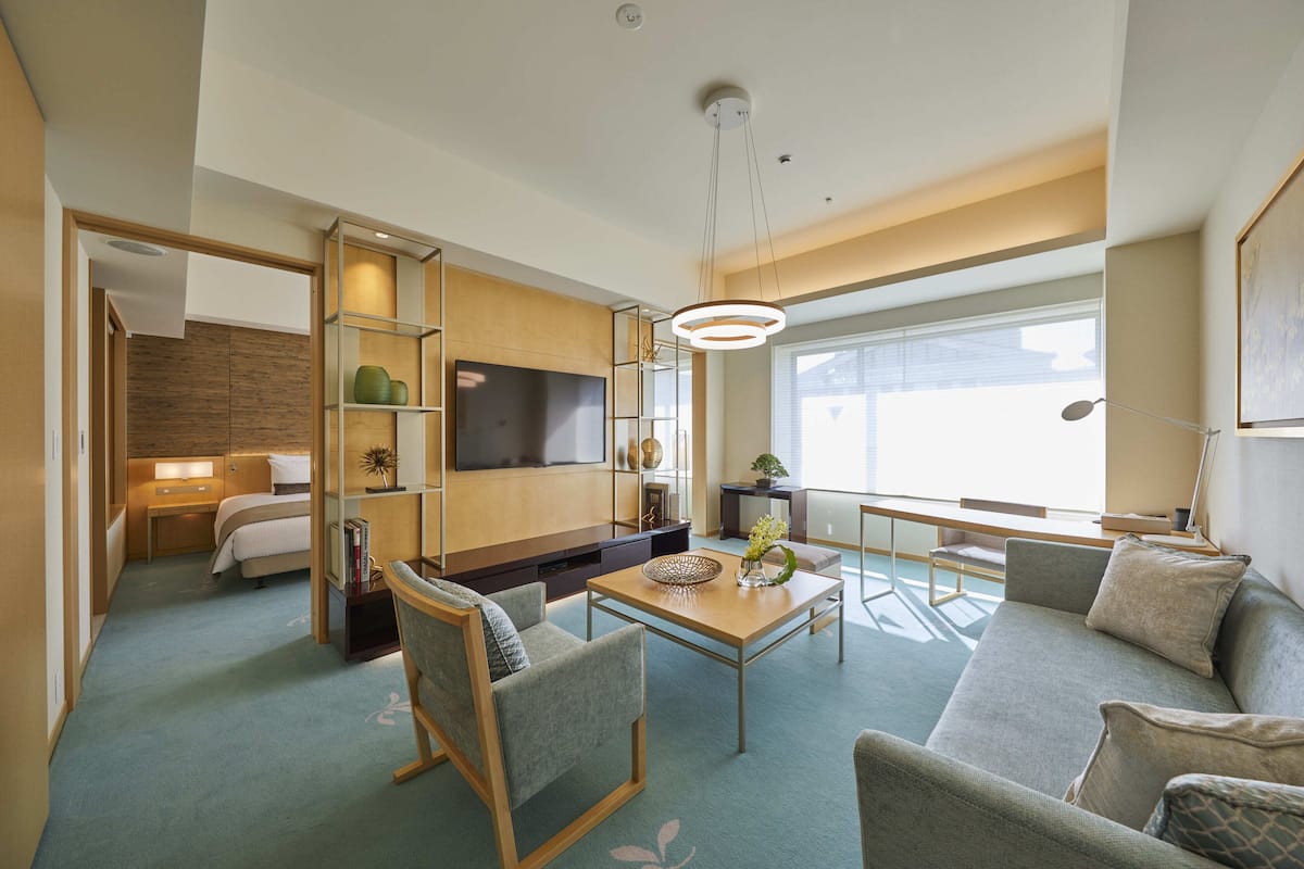 Una habitación de hotel moderna con sofá, sillones, televisor, mesa de comedor y un amplio ventanal.