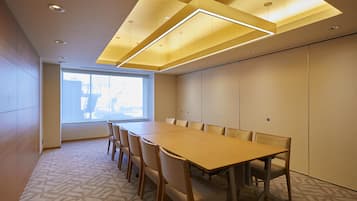 A conference room with a long table and chairs, recessed ceiling lighting, and a large window with blinds.