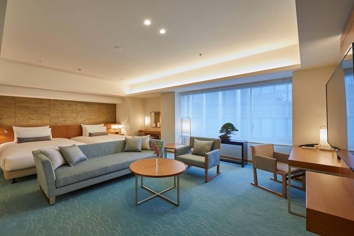 Habitación de hotel con cama, sofá, sillón, una mesita y televisor.