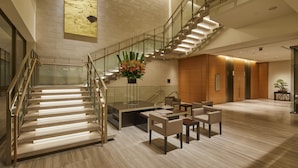 Een moderne hotel lobby met een glazen trap, een zithoek en een groot abstract schilderij aan de muur.
