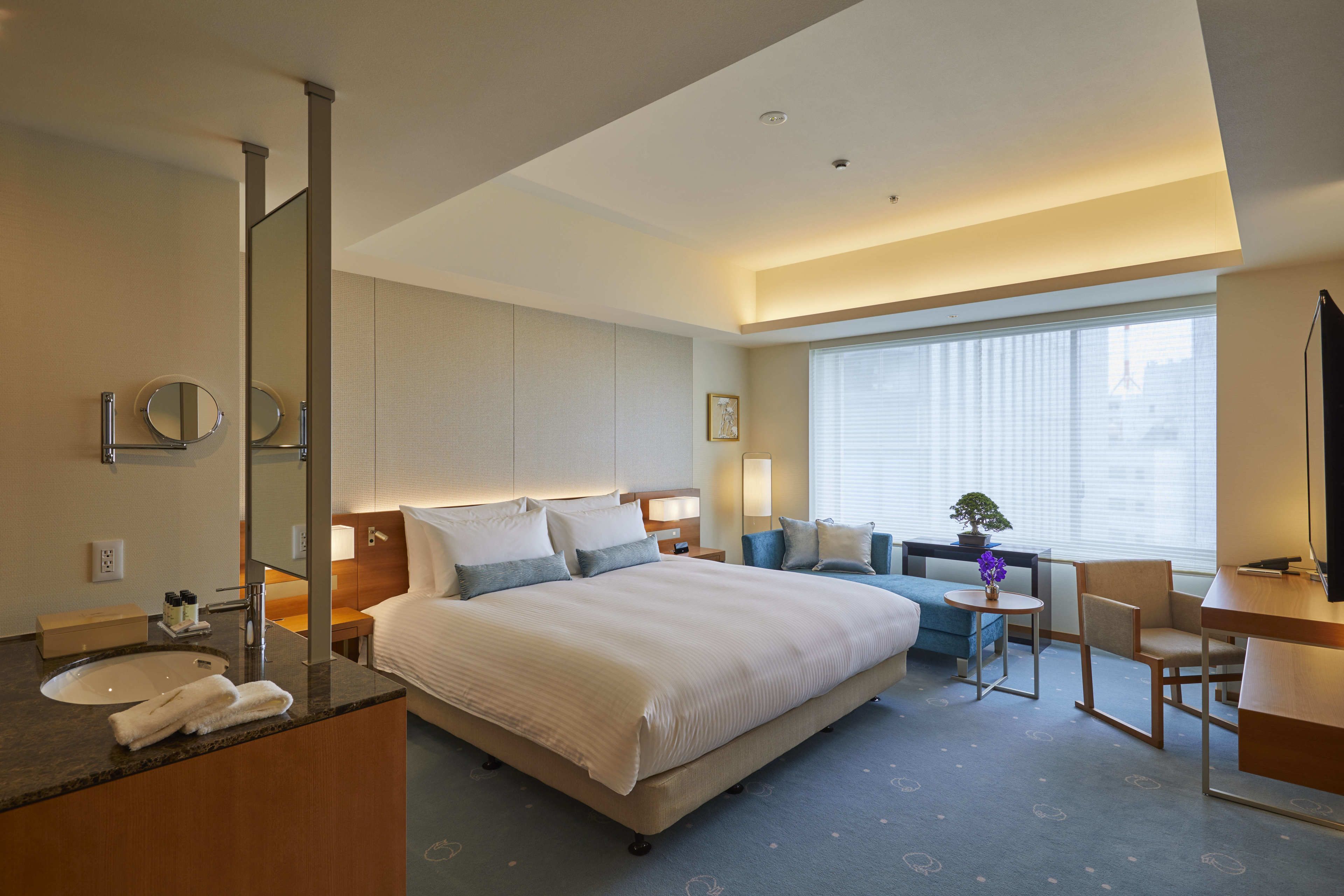 A hotel room with a large bed, a desk, a chair, a small table, a mirror, and a window with blinds.