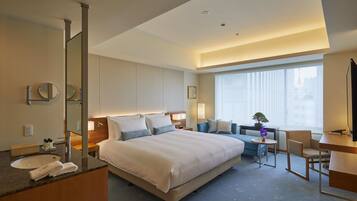 A hotel room with a large bed, a desk, a chair, a small table, a mirror, and a window with blinds.