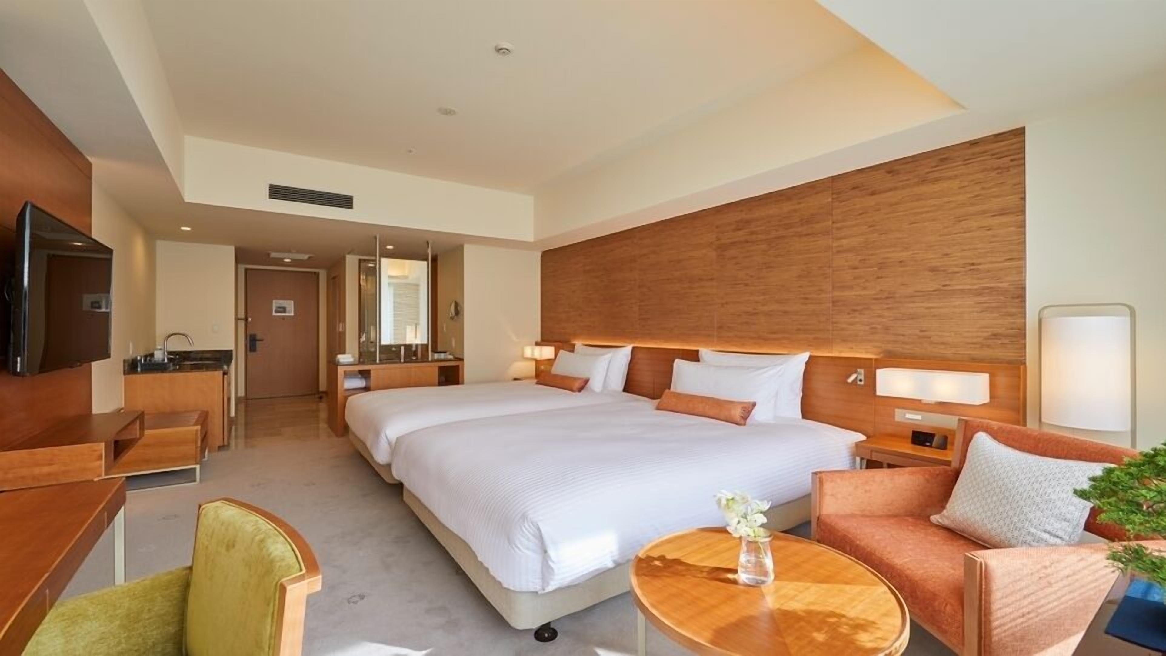 A hotel room with a large bed, a desk, a chair, a sofa, a TV, and a small table with a vase.