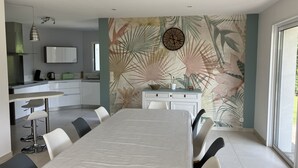 A modern kitchen with white cabinets, a dining area with a long table and chairs, and a tropical leaf-patterned wall. - House near the sea (500 m) (Le Tour-du-Parc)