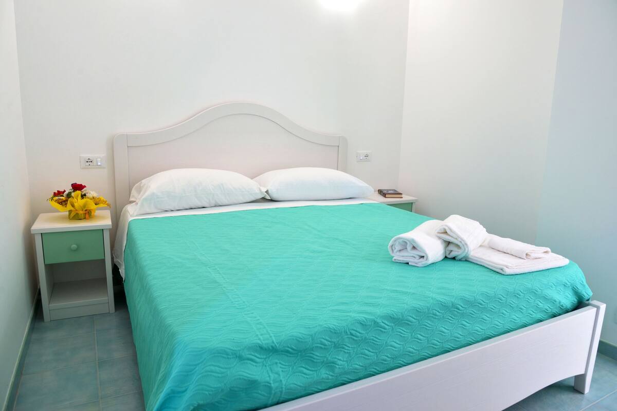 A neatly made bed with a teal bedspread, white pillows, and folded white towels on the side.