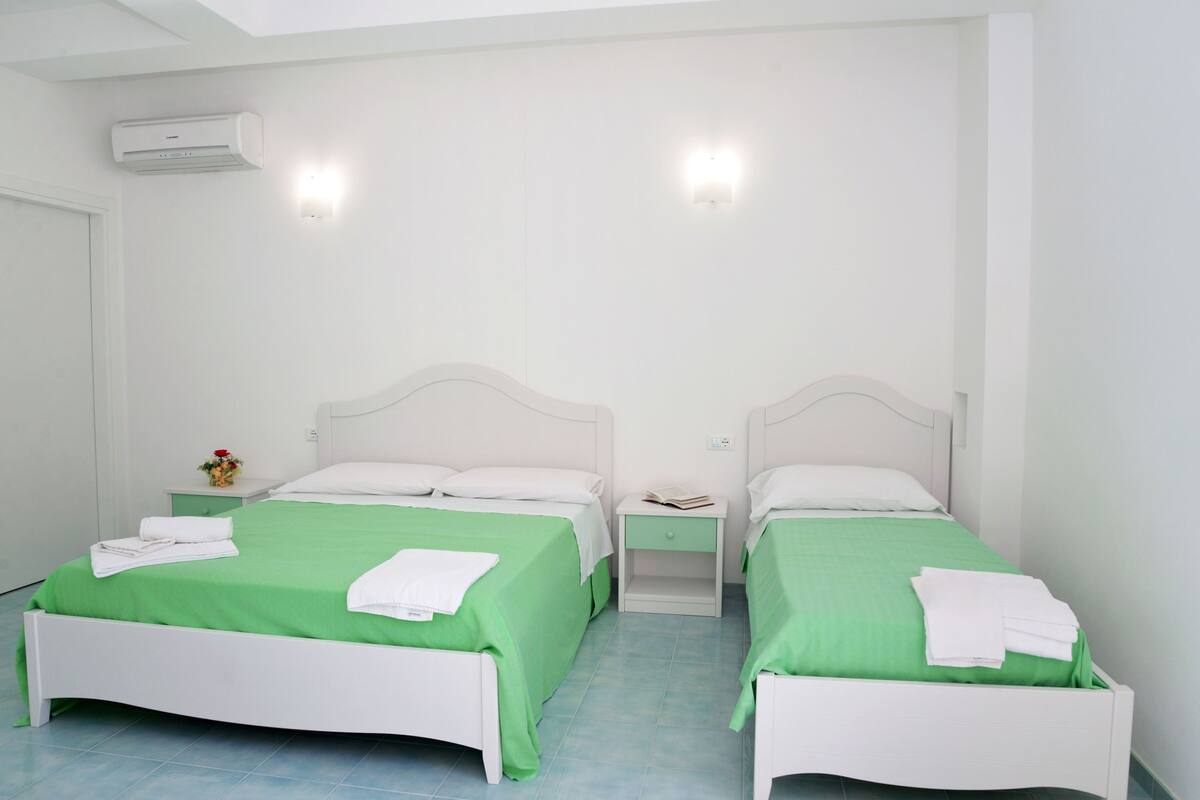 A twin room with two beds, each with a green bedspread and white pillows, a nightstand with a book and a lamp, and an air conditioning unit on the wall.
