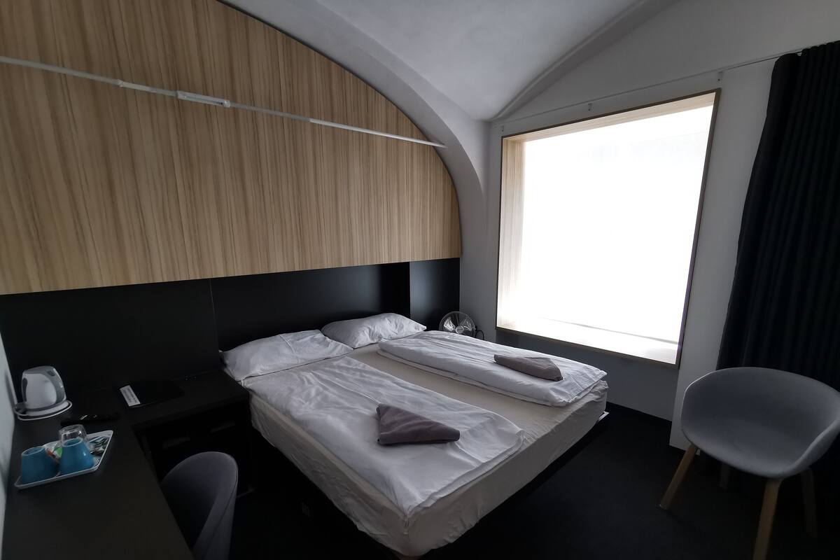 A hotel room with two beds, a desk with a kettle and cups, a chair, and a large window with curtains.