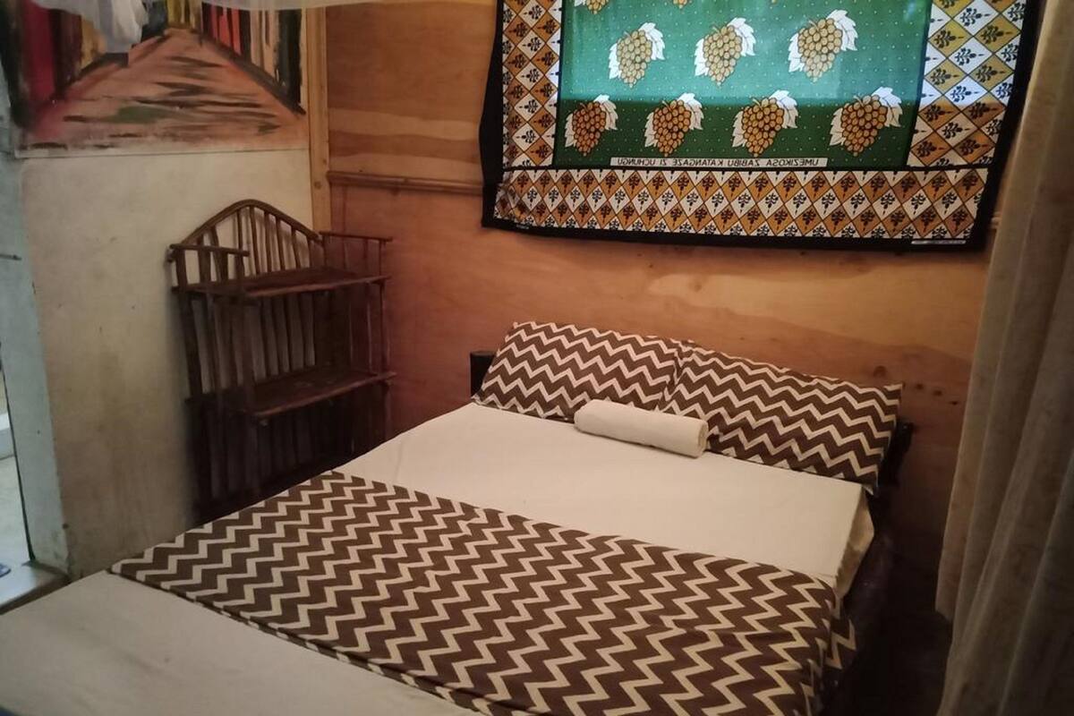 A small, cozy room with a bed, a wooden chair, and a patterned wall hanging.