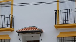 A building with a white facade, yellow trim, and a tiled roof. It features a wooden door with a green decorative panel, a small porch with a tiled roof, and a stone base. - Villa Remedios, whole house located in rural environment. (Málaga)