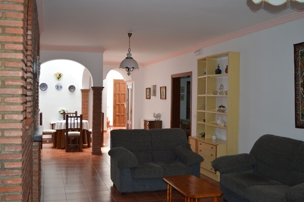 A living room with two sofas, a wooden coffee table, and a bookshelf. There is a dining area with a table and chairs in the background.