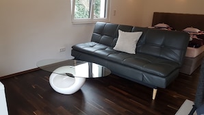 A modern living room with a dark leather sofa, a glass coffee table, and a window with a view of greenery. - Luxus Apartment Zaubersee (Bergisch Gladbach)
