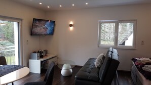 A modern living room with a black leather sofa, a glass coffee table, a flat-screen TV mounted on the wall, and a window with a view of trees. - Luxus Apartment Zaubersee (Bergisch Gladbach)