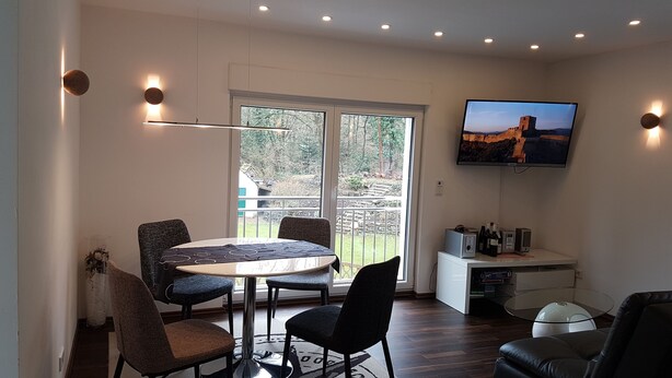 A modern living room with a round table, four chairs, a flat-screen TV mounted on the wall, and a small entertainment unit with speakers and a gaming console. - Luxus Apartment Zaubersee (Bergisch Gladbach)