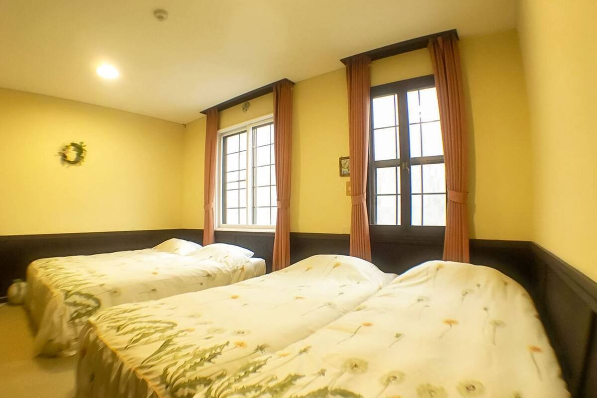 Ein Hotelzimmer mit zwei Einzelbetten, einem Fenster mit Vorhängen, einer Wanduhr und einem Kranz an der Wand.